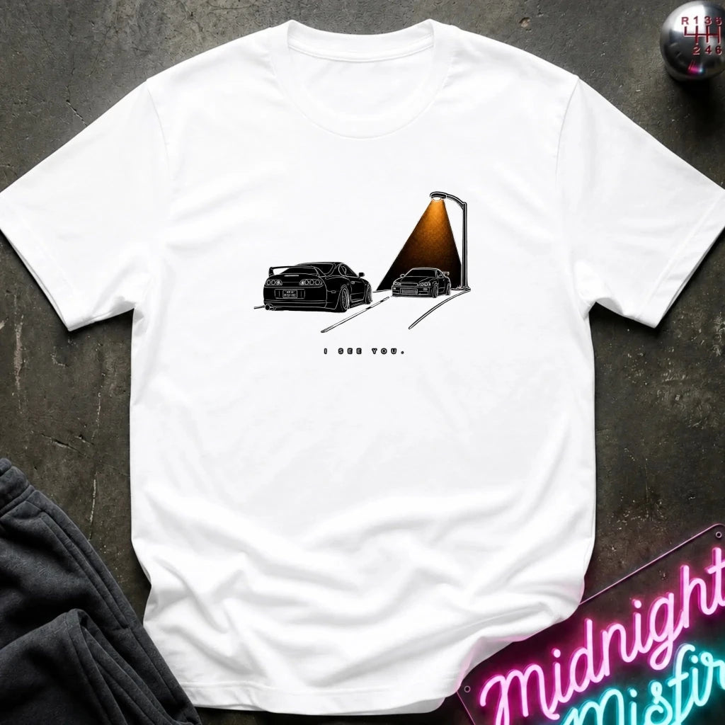 A white short-sleeve t-shirt laid flat on a dark textured surface, featuring a graphic print of two black sports cars passing each other.