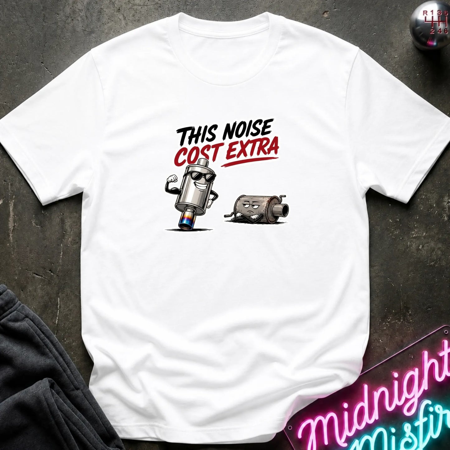 A white short-sleeve graphic T-shirt laid flat on a dark textured surface. The shirt features with the text 'THIS NOISE COST EXTRA' in black and red.