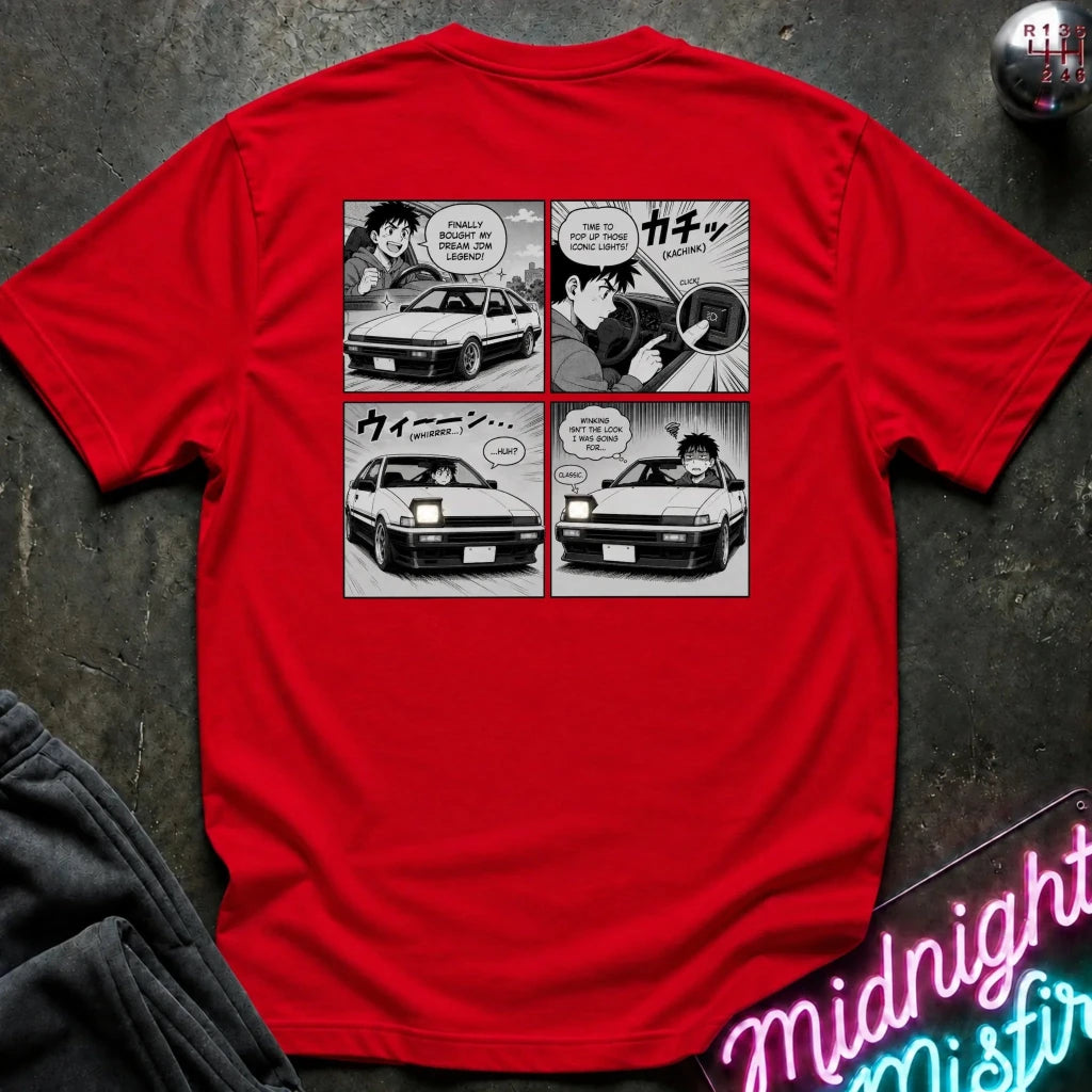 A red short-sleeve t-shirt laid flat on a dark textured surface, showing the back side with a black-and-white four-panel comic print featuring a car and Japanese text. A neon sign and dark fabric are partially visible around it.