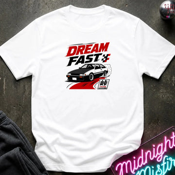 A white short-sleeve graphic T-shirt laid flat on a dark textured surface. The shirt features a red, black, and white 'DREAM FAST' graphic with a car illustration.