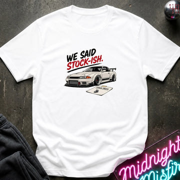 A white short-sleeve graphic T-shirt laid flat on a dark textured surface. The shirt features a printed design of a white sports car with the text 'WE SAID STOCK-ISH.