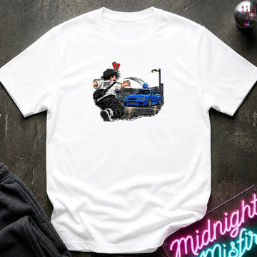 A white short-sleeve crewneck t-shirt laid flat on a dark textured surface. The center of the shirt features a graphic print of an anime character in a black and white outfit with a hat, next to a blue sports car under a streetlamp.