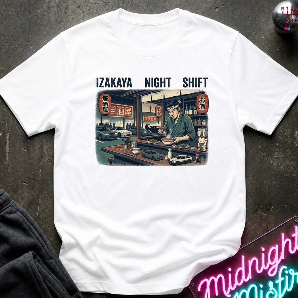 A white short-sleeve t-shirt laid flat on a dark textured surface, featuring a vintage-style graphic of an izakaya scene with the text 'IZAKAYA NIGHT SHIFT' above it. A neon sign and dark clothing are partially visible in the styled flat-lay composition.