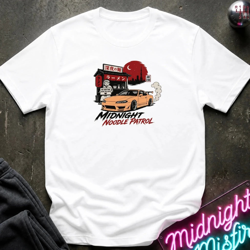 white crewneck t-shirt featuring a graphic print of a yellow Nissan Silvia S15 drift car in front of a red moon and Japanese storefront, with the text 'MIDNIGHT Noodle PATROL'. The shirt is laid flat on a dark textured surface with a neon sign and dark pants partially visible.