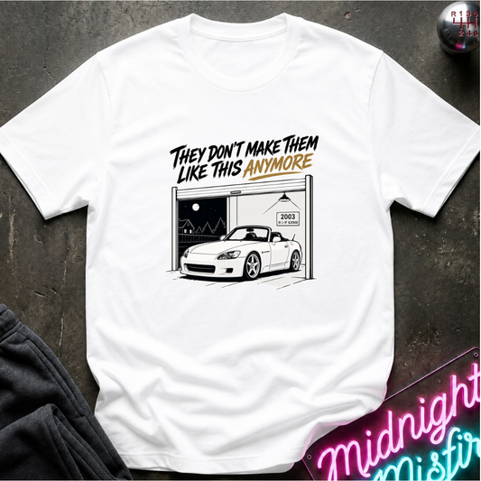 A white short-sleeve graphic t-shirt laid flat on a dark textured surface. The graphic features the text 'THEY DON'T MAKE THEM LIKE THIS ANYMORE' in black and orange lettering above a black-and-white line drawing of a white convertible sports car parked in a garage.