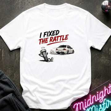 A white short-sleeve graphic T-shirt laid flat on a dark textured surface. The shirt features a printed design with the text 'I FIXED THE RATTLE' in black and red, above a cartoon character and a white sports car.
