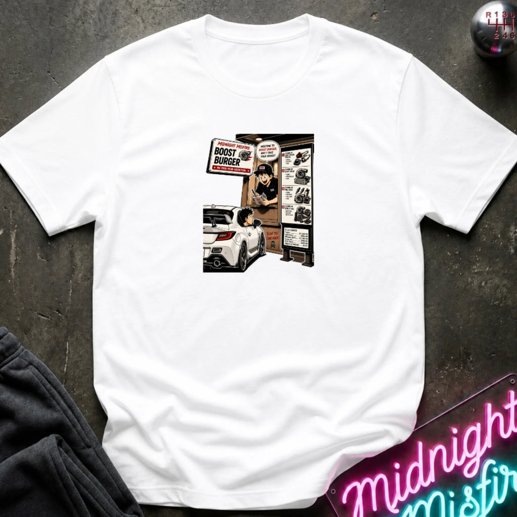 A white short-sleeve crewneck t-shirt laid flat on a dark textured surface, featuring a central graphic print of a street-food burger scene with a white car and signs. 