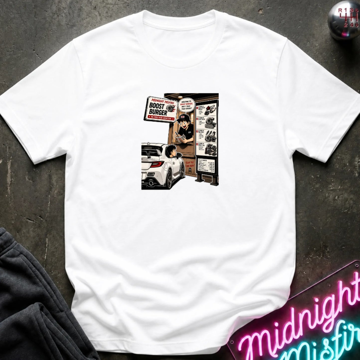 A white short-sleeve crewneck t-shirt laid flat on a dark textured surface, featuring a central graphic print of a street-food burger scene with a white car and signs. 