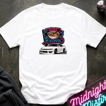 A white short-sleeve graphic t-shirt laid flat on a dark textured surface. The graphic features a white sports car beneath a ramen bowl illustration with neon-style text reading 'Ramen & Drift Diner' and Japanese characters.