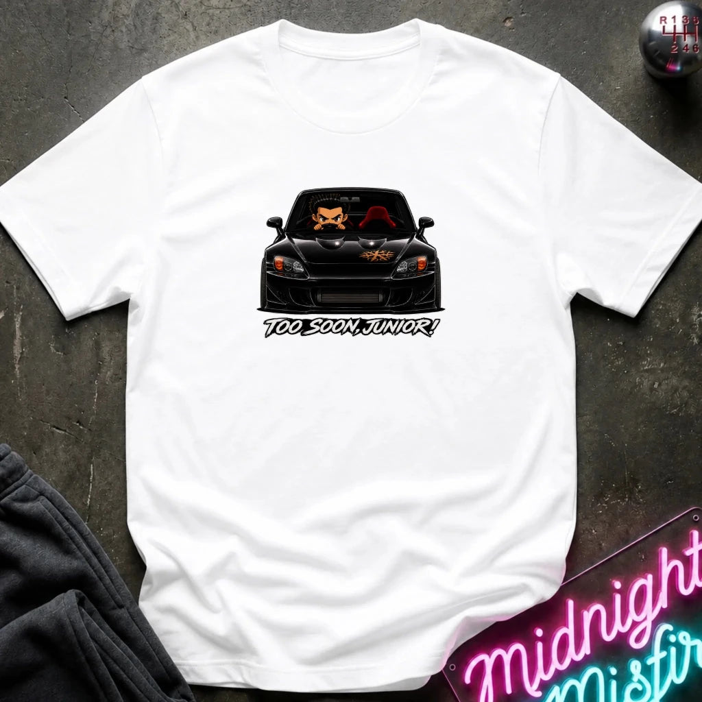 A white short-sleeve t-shirt laid flat on a dark textured surface, featuring a graphic of a black sports car with orange and red details and the text 'TOo SOON, JUNIOR!' printed below it.