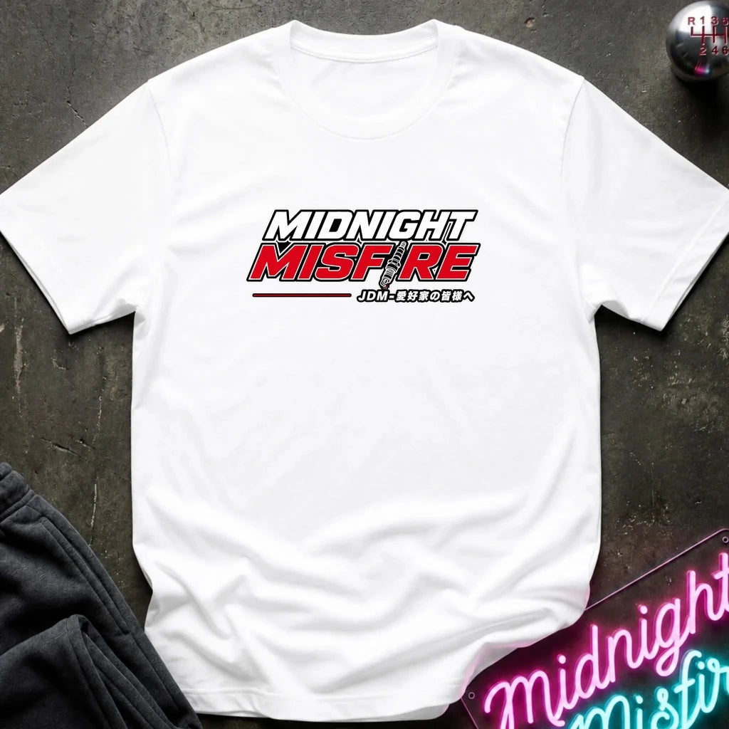 A white short-sleeve t-shirt with a 'Midnight Misfire' graphic printed on the chest, laid flat on a dark textured surface with a neon sign and dark pants partially visible.