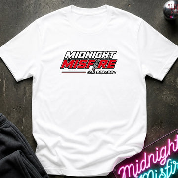 A white short-sleeve t-shirt with a 'Midnight Misfire' graphic printed on the chest, laid flat on a dark textured surface with a neon sign and dark pants partially visible.