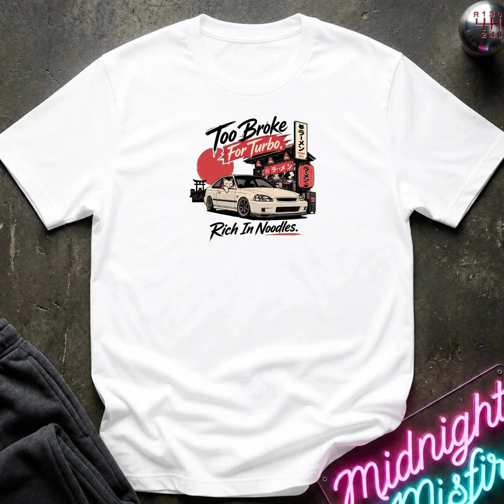 A white short-sleeve graphic t-shirt laid flat on a dark textured surface. The graphic features a white sports car in front of a red and black Japanese-style street sign with the text 'Too Broke To For Turbo.