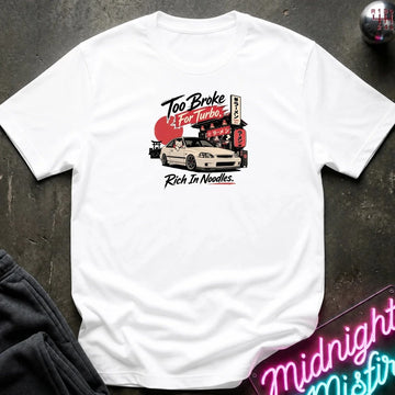 A white short-sleeve graphic t-shirt laid flat on a dark textured surface. The graphic features a white sports car in front of a red and black Japanese-style street sign with the text 'Too Broke To For Turbo.