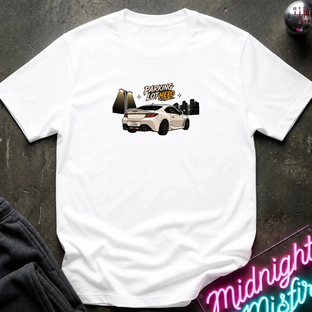 A white crewneck t-shirt laid flat on a dark textured surface, featuring a graphic of a white sports car with the text 'PARKING LOT HERO'