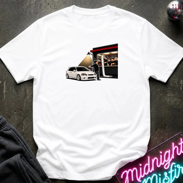 A white short-sleeve crewneck t-shirt laid flat on a dark textured surface. The shirt features a rectangular graphic print on the chest showing a white hatchback car parked in front of a building with a red awning and a person standing by the door.