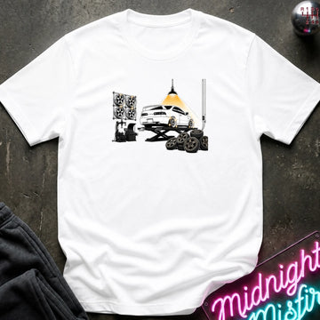 A white short-sleeve crewneck t-shirt laid flat on a dark textured surface. The shirt features a graphic print of a white car on a hydraulic lift with new wheels.