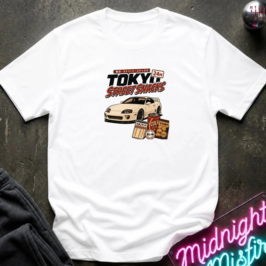 white  short-sleeve graphic T-shirt laid flat on a dark textured surface. The shirt features a central graphic with the text 'Tokyo Street Snacks' above a white sports car and snack illustrations.