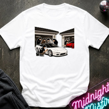 A white short-sleeve crewneck t-shirt featuring a graphic print of a white sports car and several anime characters in front of a house. The shirt is displayed flat on a dark textured surface with a neon sign and dark sweatpants partially visible.