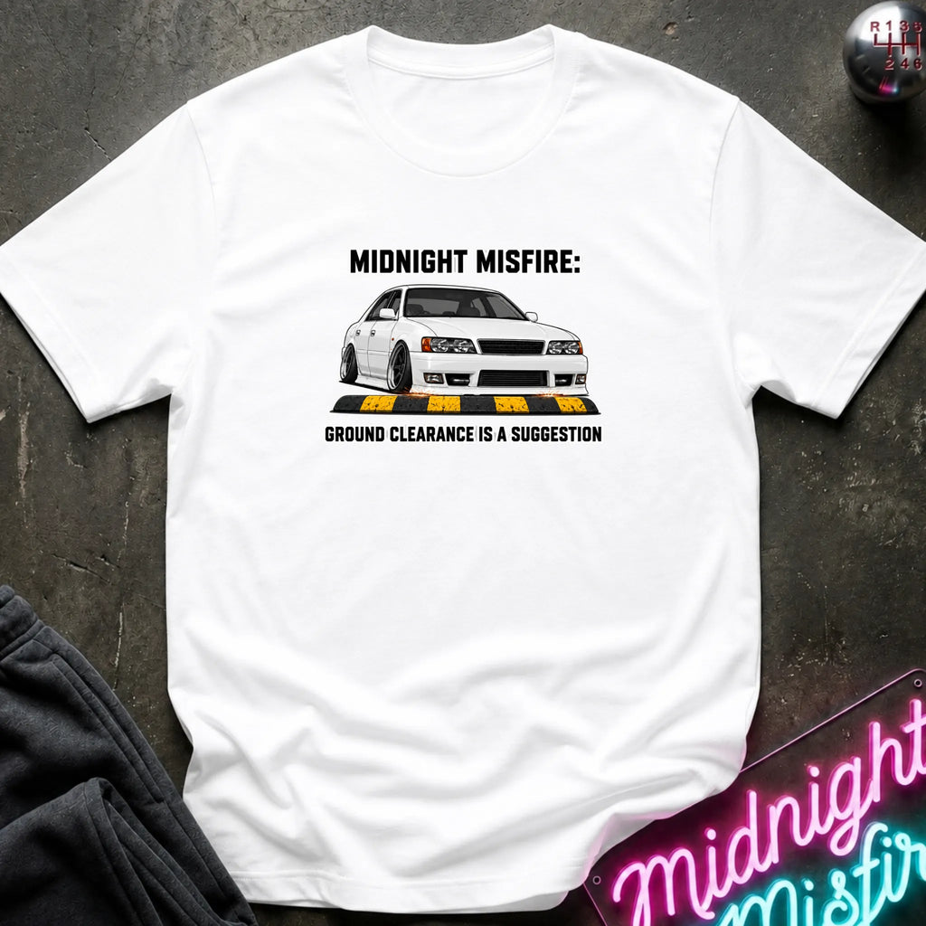 A white short-sleeve graphic t-shirt laid flat on a dark textured surface. The shirt features a printed graphic of a white sports car with black text reading 'MIDNIGHT MISFIRE: GROUND CLEARANCE IS A SUGGESTION'.