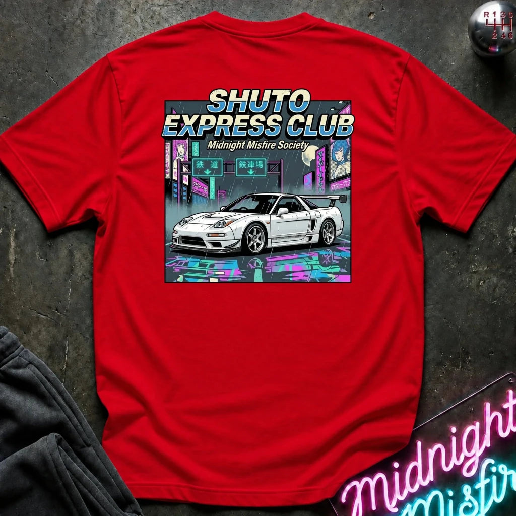 A red short-sleeve t-shirt laid flat on a dark textured surface, featuring a large graphic print on the back with the text 'SHUTO EXPRESS CLUB' and 'Midnight Misfire Society' above an illustration of a white sports car in a neon city scene. A gear shift knob and a neon sign are partially visible as props.