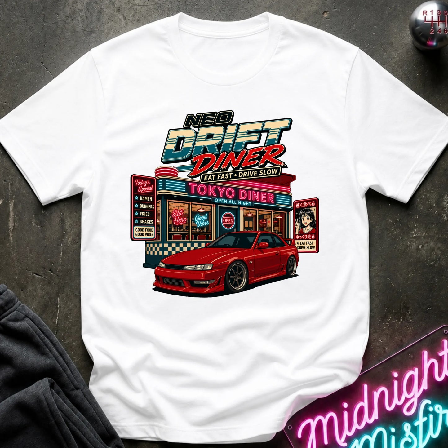 A white short-sleeve graphic T-shirt laid flat on a dark textured surface. The shirt features a colorful retro diner illustration with the text 'NEO DRIFT DINER' and 'TOKYO DINER,' plus a red sports car graphic.