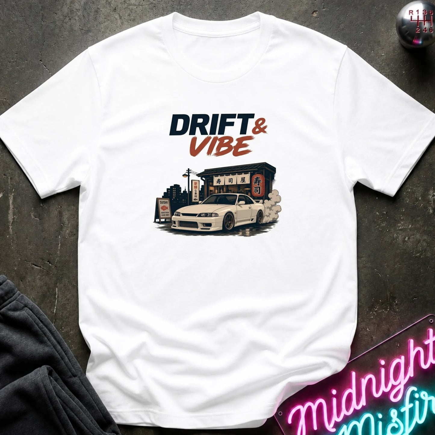 A white short-sleeve graphic T-shirt laid flat on a dark textured surface. The shirt features a printed design with the text 'DRIFT & VIBE' above an illustration of a white sports car in front of a building.