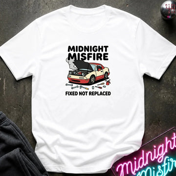 A white short-sleeve graphic T-shirt laid flat on a dark textured surface. The shirt features a printed design of a beige and red car with its hood open, accompanied by tools and the text 'MIDNIGHT MISFIRE' and 'FIXED NOT REPLACED'.
