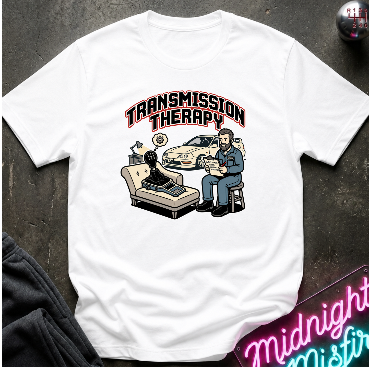 A white short-sleeve graphic t-shirt laid flat on a dark textured surface. The graphic features the text 'TRANSMISSION THERAPY' above an illustration of a therapist sitting on a stool with a patient in a chair, alongside a white car.