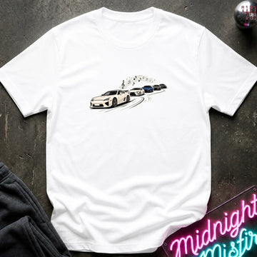 A white short-sleeve crewneck t-shirt featuring a graphic print of a white sports car leading a line of other cars, laid flat on a dark textured surface with a neon sign and dark pants partially visible.