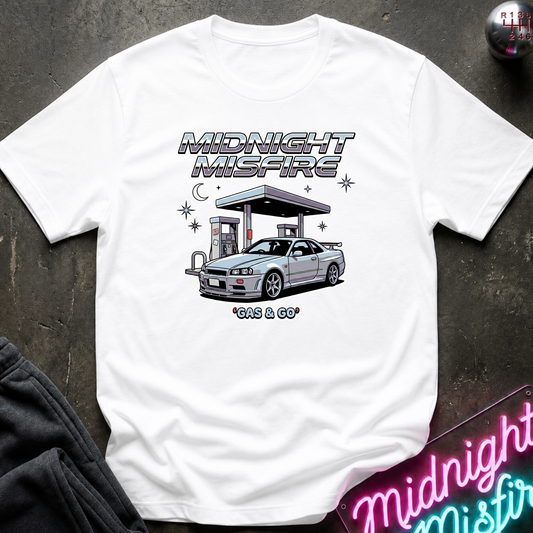 A white short-sleeve graphic T-shirt laid flat on a dark textured surface. The shirt features a printed illustration of a silver sports car at a gas station with the text 'MIDNIGHT MISFIRE' and '*GAS & GO*' beneath it.