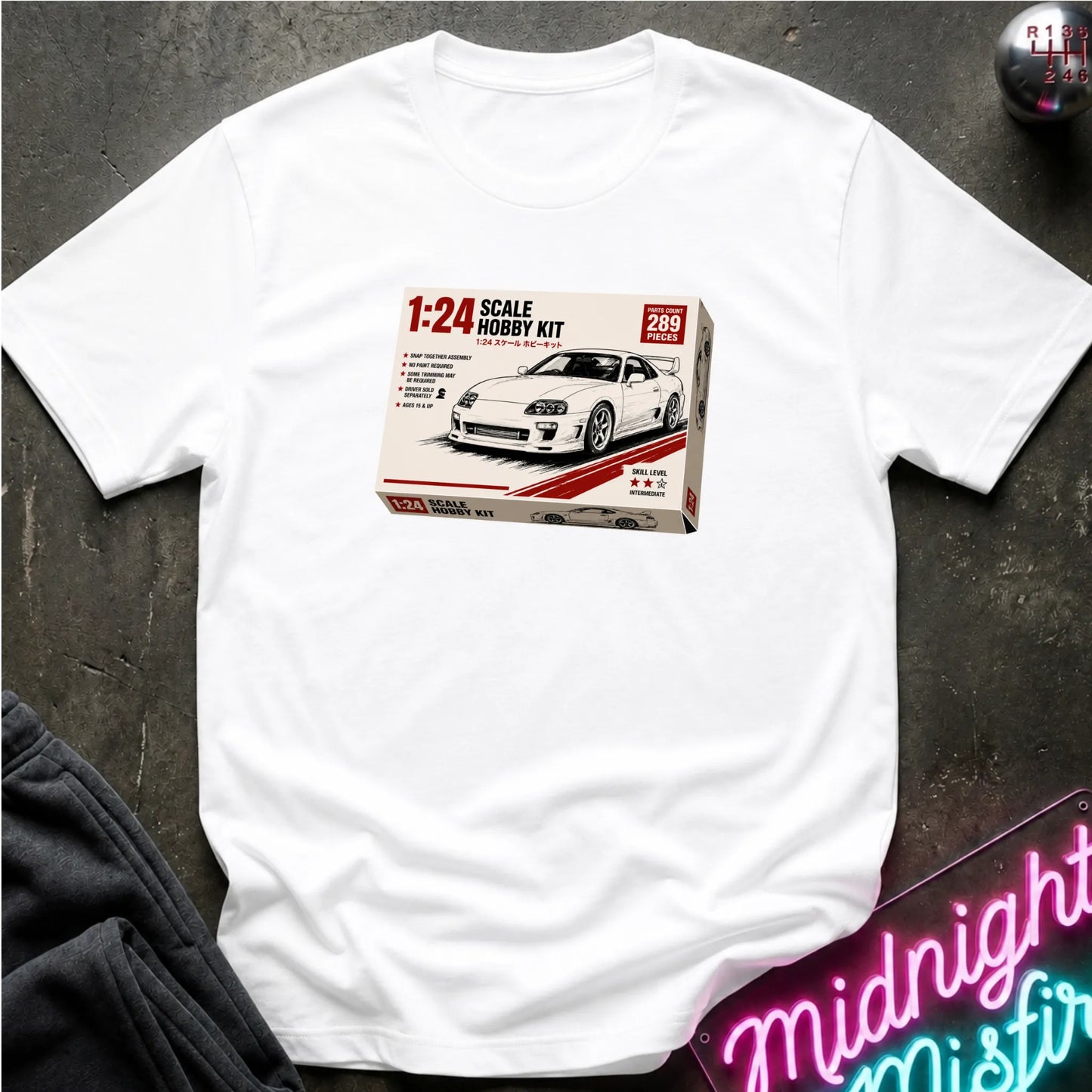A white short-sleeve crewneck t-shirt laid flat on a dark textured surface, featuring a graphic print of a 1:24 scale model kit box for a white sports car. The shirt is styled with a dark sweatshirt in the corner and a neon sign in the bottom right.