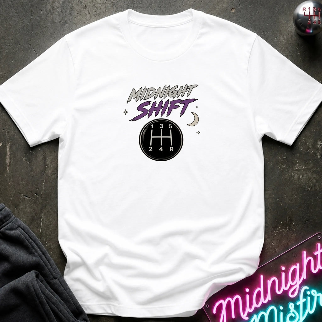 A white short-sleeve graphic t-shirt laid flat on a dark textured surface. The graphic features the words 'MIDNIGHT SHIFT' in stylized gray and purple lettering above a black circular gear shift pattern.