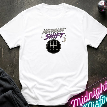 A white short-sleeve graphic t-shirt laid flat on a dark textured surface. The graphic features the words 'MIDNIGHT SHIFT' in stylized gray and purple lettering above a black circular gear shift pattern.