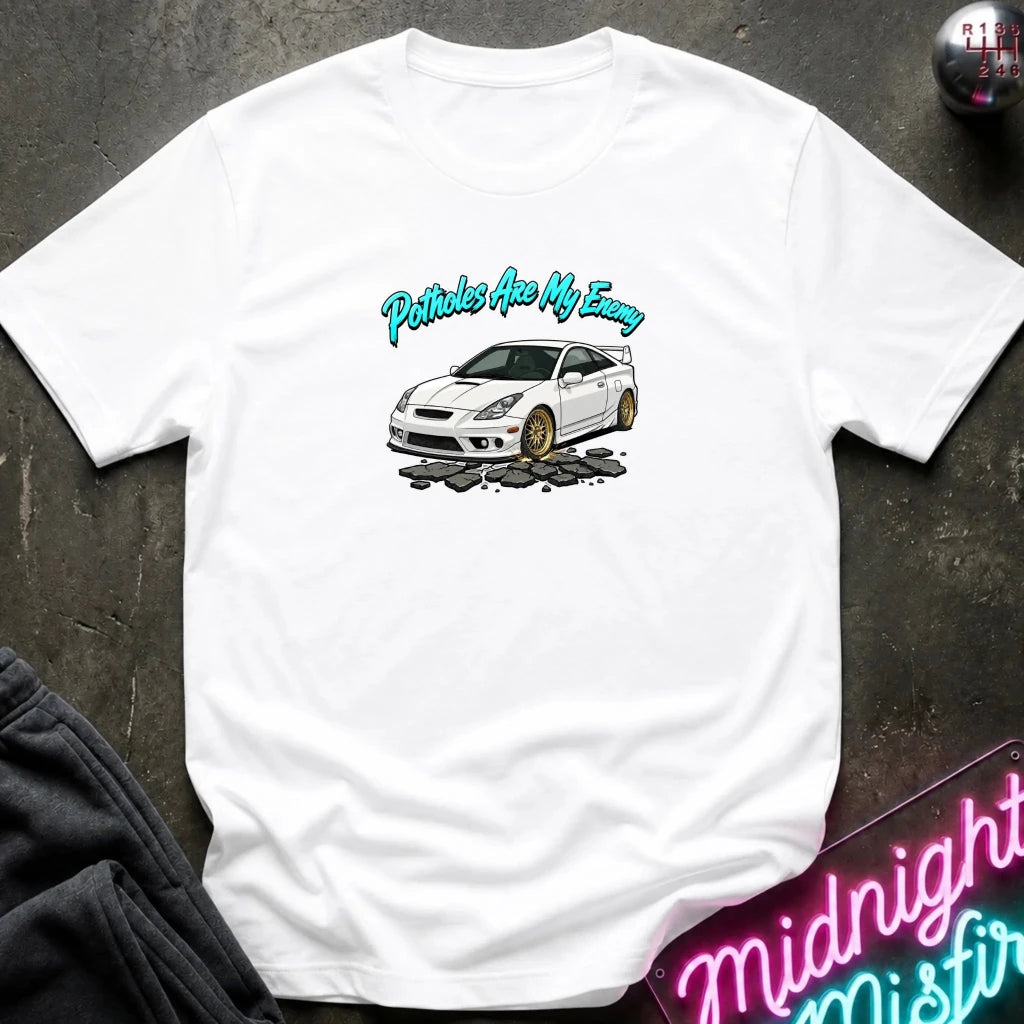 A white short-sleeve t-shirt laid flat on a dark textured surface, featuring a graphic of a white sports car with gold wheels and the text 'Petrols Are My Enemy' in teal. A neon sign and dark clothing are partially visible in the corners.