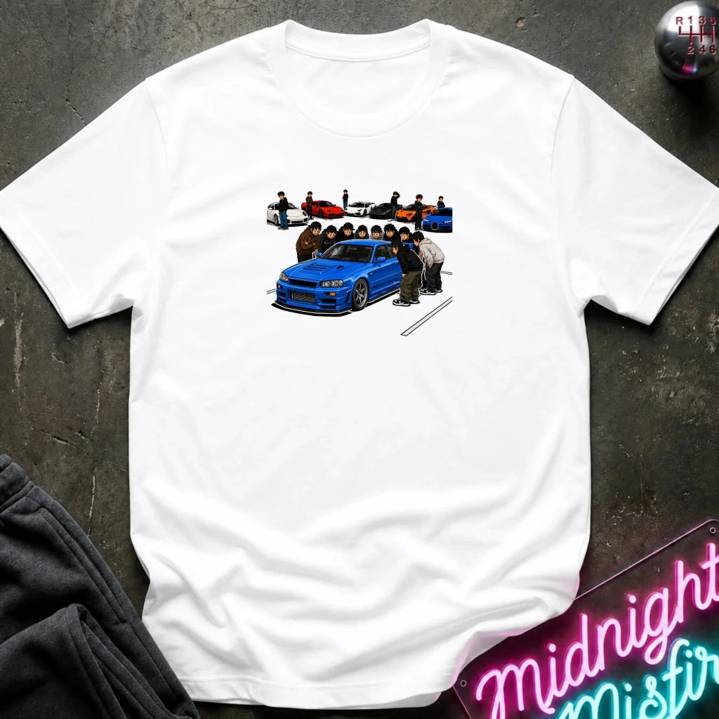 A white short-sleeve crewneck t-shirt laid flat on a dark textured surface. The shirt features a graphic print in the center depicting a blue sports car with several people standing around it, and a small row of colorful cars above.