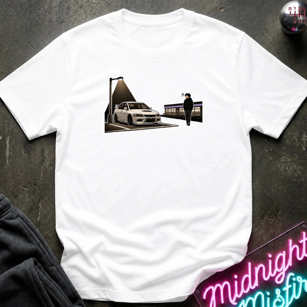 A white short-sleeve t-shirt laid flat on a dark textured surface, featuring a graphic print of a white sports car under a streetlamp with a figure standing nearby. Partially visible props include dark pants in the lower-left corner and a neon sign in the lower-right.