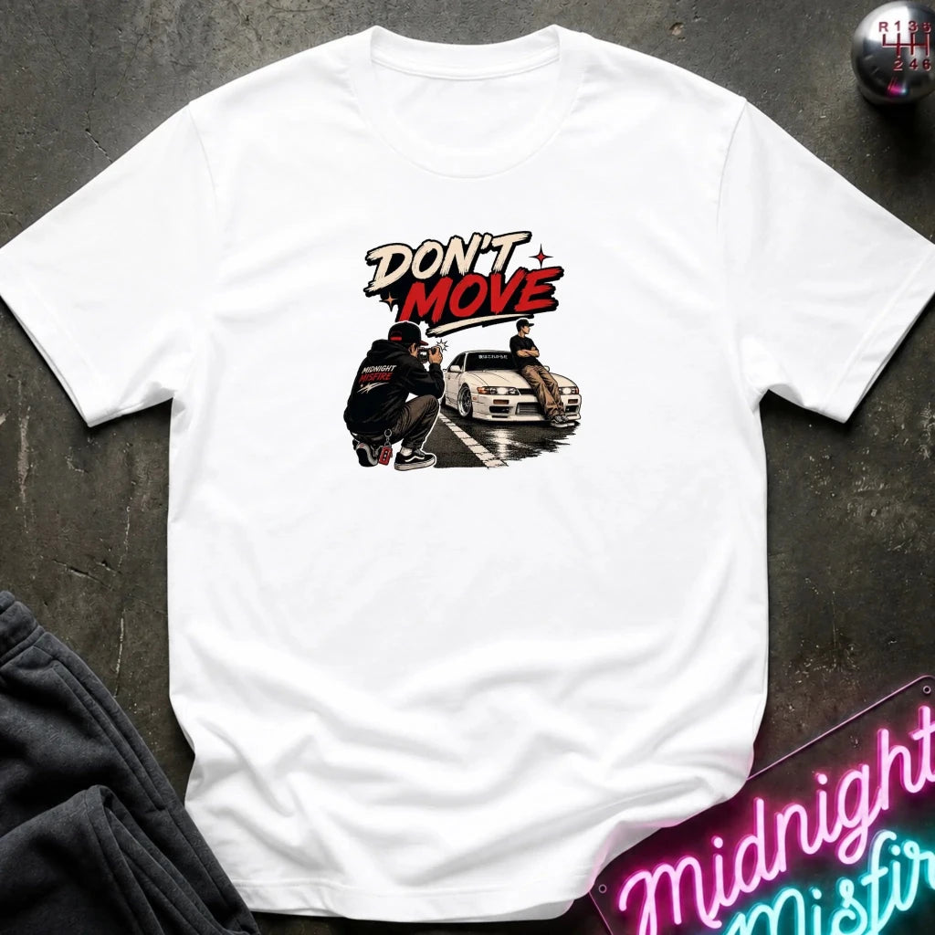 A white short-sleeve graphic t-shirt laid flat on a dark textured surface. The graphic features the text 'DON'T MOVE' in stylized red and black lettering above an illustration of a person crouching next to a white car.