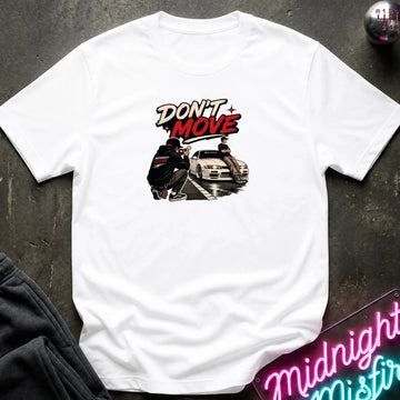 A white short-sleeve graphic t-shirt laid flat on a dark textured surface. The graphic features the text 'DON'T MOVE' in stylized red and black lettering above an illustration of a person crouching next to a white car.