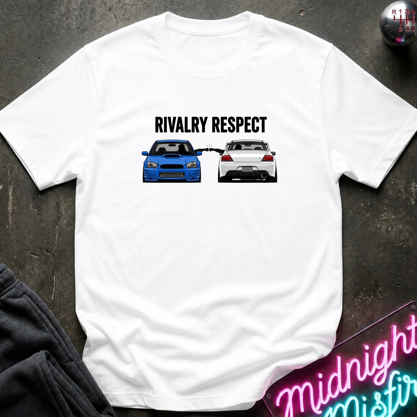 A white short-sleeve graphic t-shirt laid flat on a dark textured surface. The graphic features the text 'RIVALRY RESPECT' above a blue and white car, 