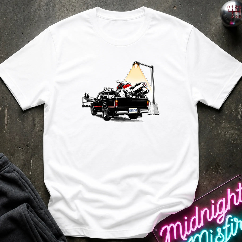 A white short-sleeve crewneck t-shirt featuring a graphic print of a black pickup truck carrying a white and red motorcycle under a street lamp, laid flat on a dark textured surface.