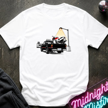 A white short-sleeve crewneck t-shirt featuring a graphic print of a black pickup truck carrying a white and red motorcycle under a street lamp, laid flat on a dark textured surface.