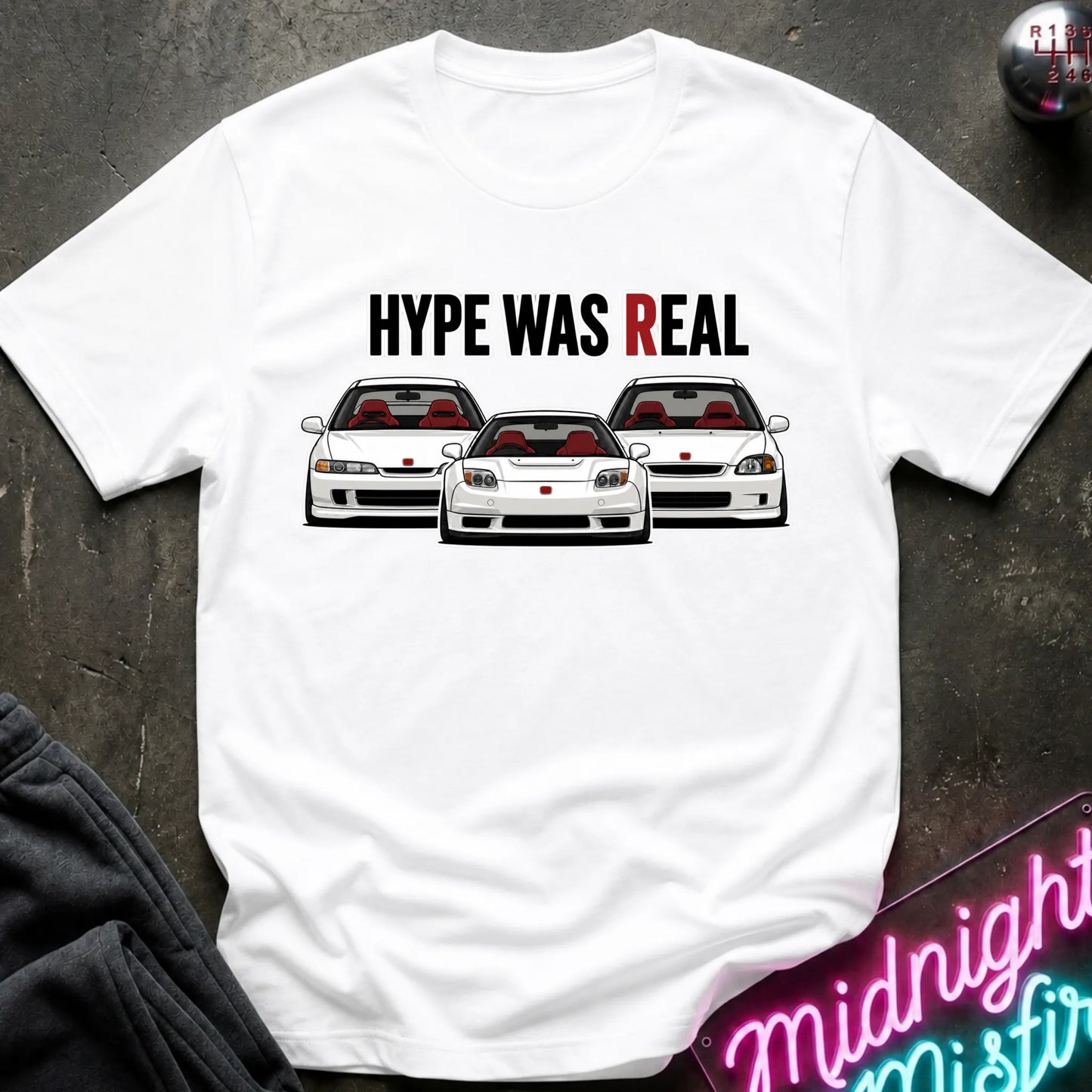 A white short-sleeve graphic T-shirt laid flat on a dark textured surface. The shirt features the text 'HYPE WAS REAL' above an illustration of three white sports cars with red interiors.