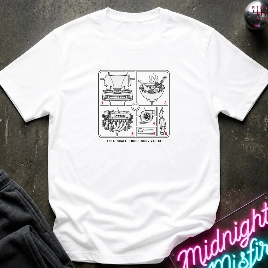 A white short-sleeve t-shirt laid flat on a dark textured surface, featuring a black and white schematic graphic of a 1:24 scale house survival kit with numbered steps