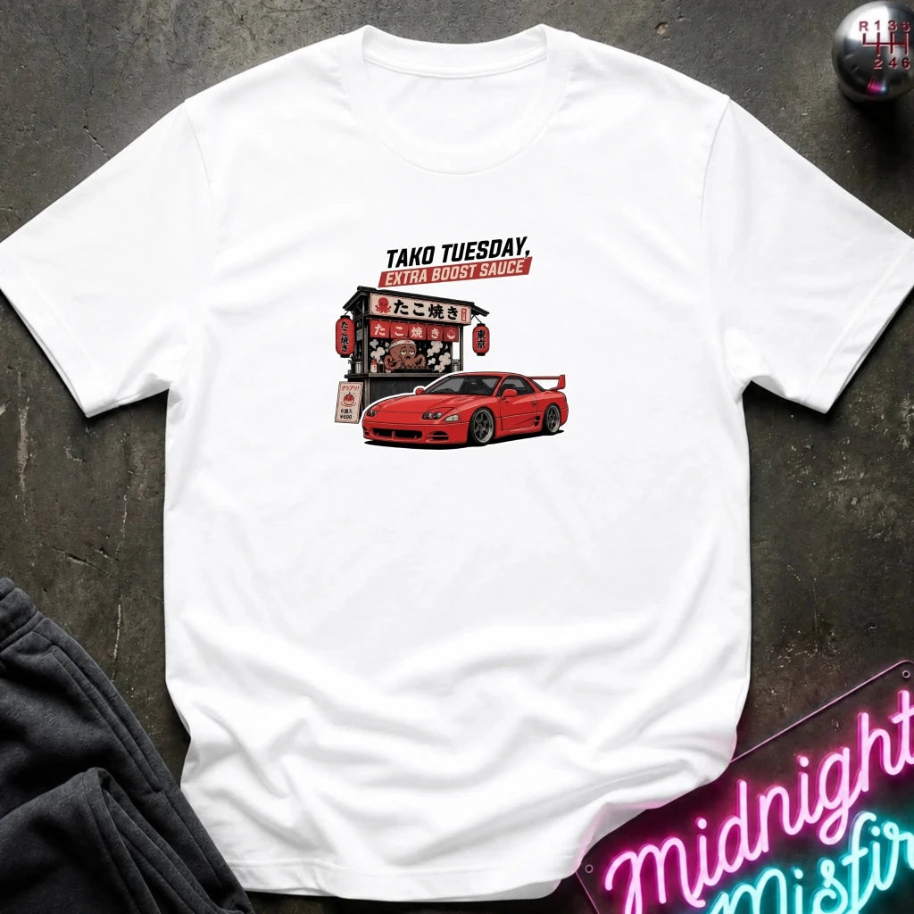 A white short-sleeve t-shirt laid flat on a dark textured surface, featuring a graphic print of a red sports car and a Japanese-style restaurant sign with the text 'TAKO TUESDAY' 