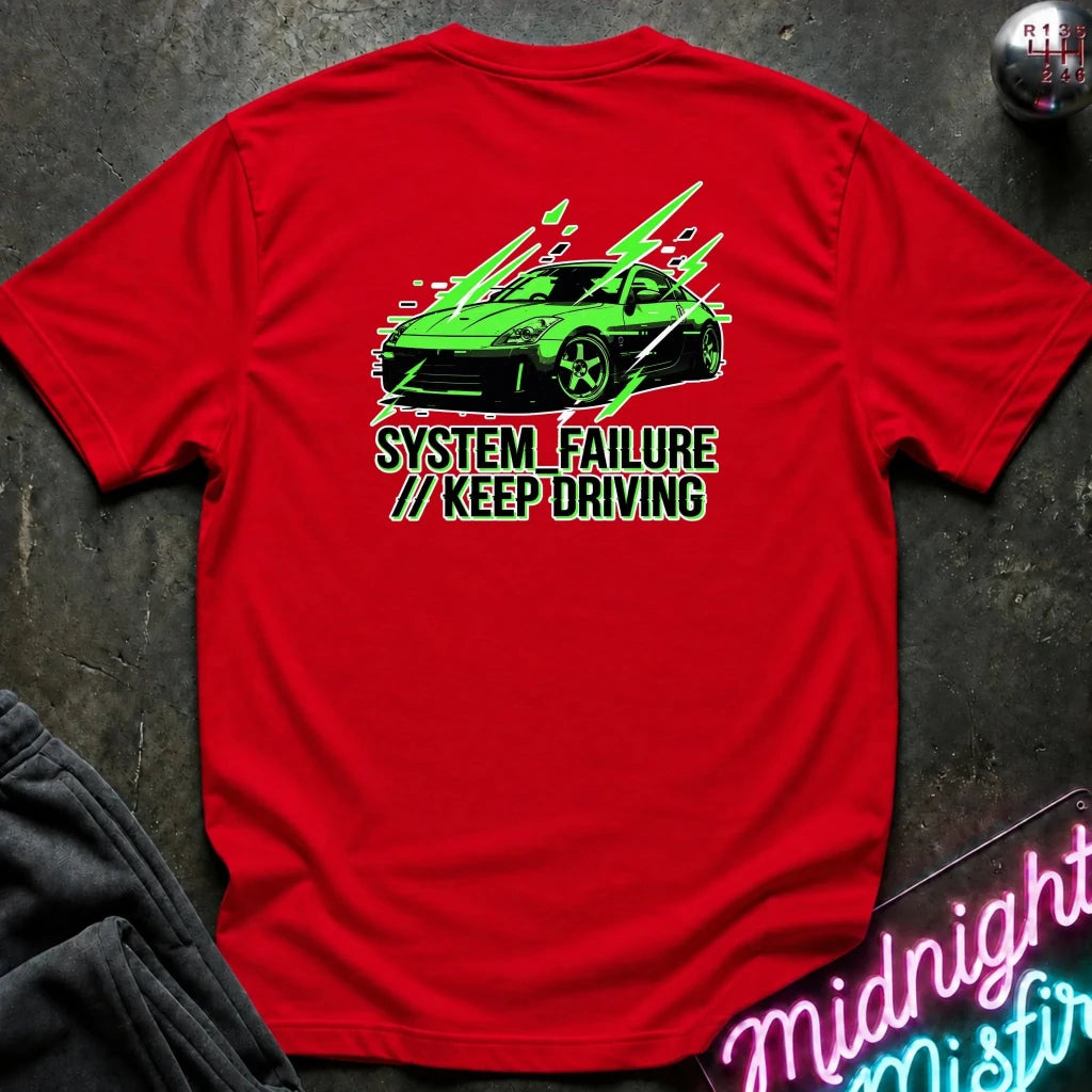 red short-sleeve t-shirt laid flat on a dark textured surface, featuring a graphic of a green sports car with text 'SYSTEM FAILURE // KEEP DRIVING' in green. A neon sign and dark clothing are partially visible as props.