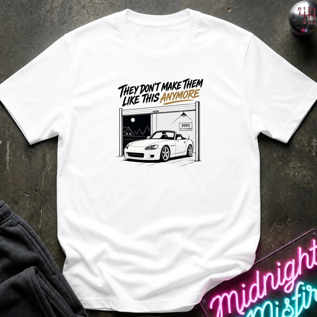 A white short-sleeve graphic t-shirt laid flat on a dark textured surface. The graphic features a black and white illustration of a white sports car parked inside a garage with the text 'THEY DON'T MAKE THEM LIKE THIS ANYMORE' in black and orange lettering.