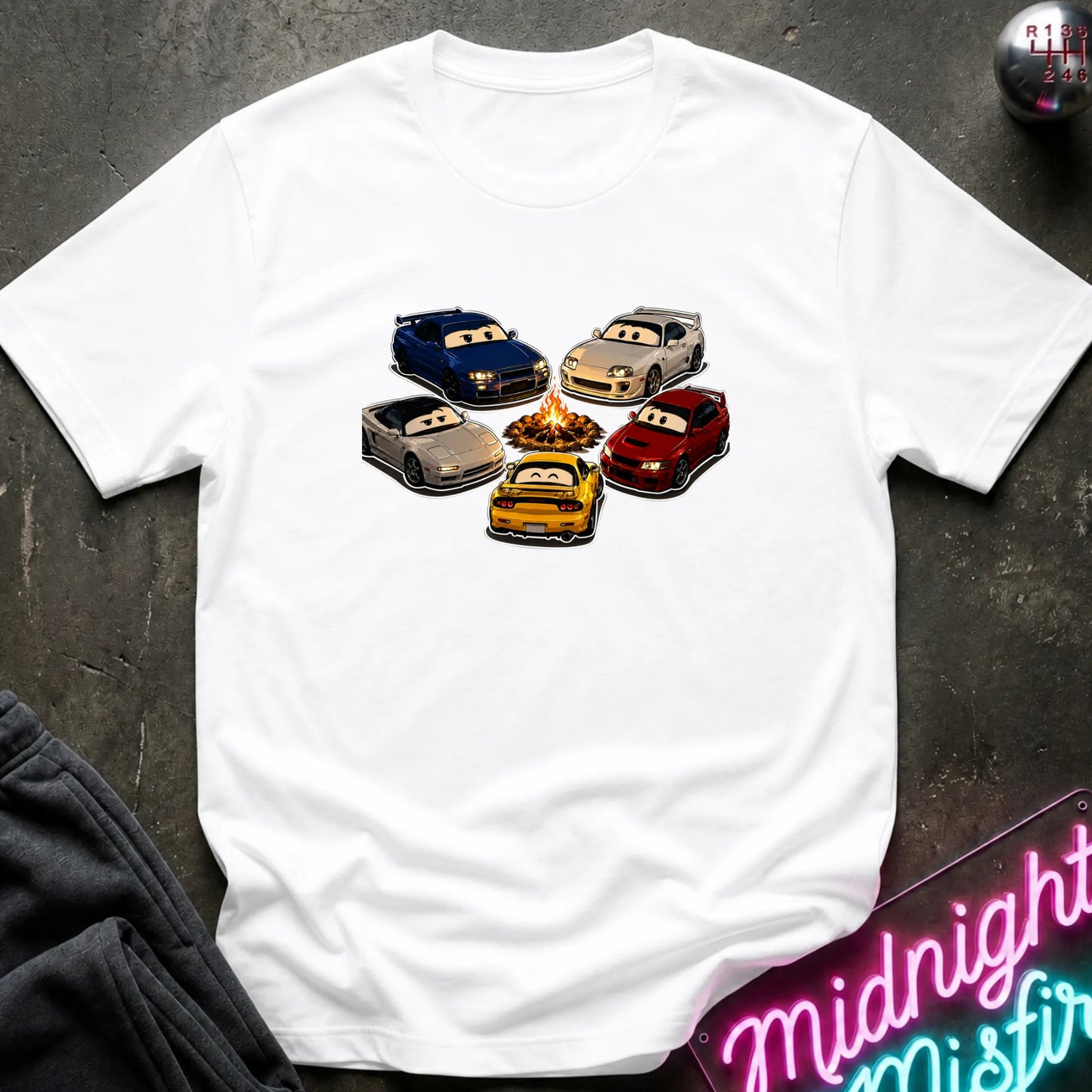 A white short-sleeve t-shirt laid flat on a dark textured surface, featuring a graphic of cartoon race cars arranged around a campfire. The shirt is styled with a gray sweatshirt in the corner and a pink neon sign in the bottom right.