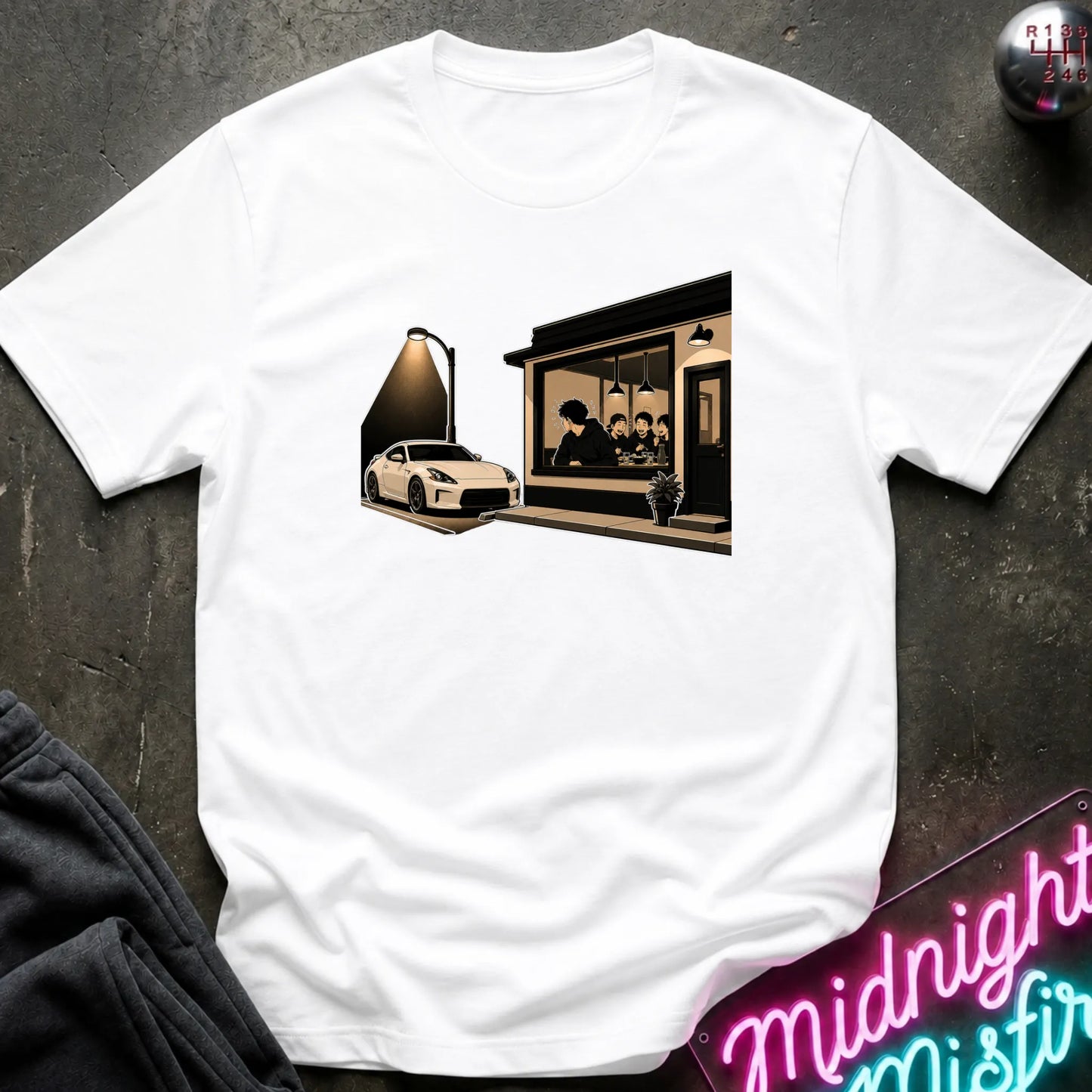 A white short-sleeve crewneck t-shirt laid flat on a dark textured surface. The center of the shirt features a graphic print of a white sports car parked outside a dark cafe with a spotlight effect.