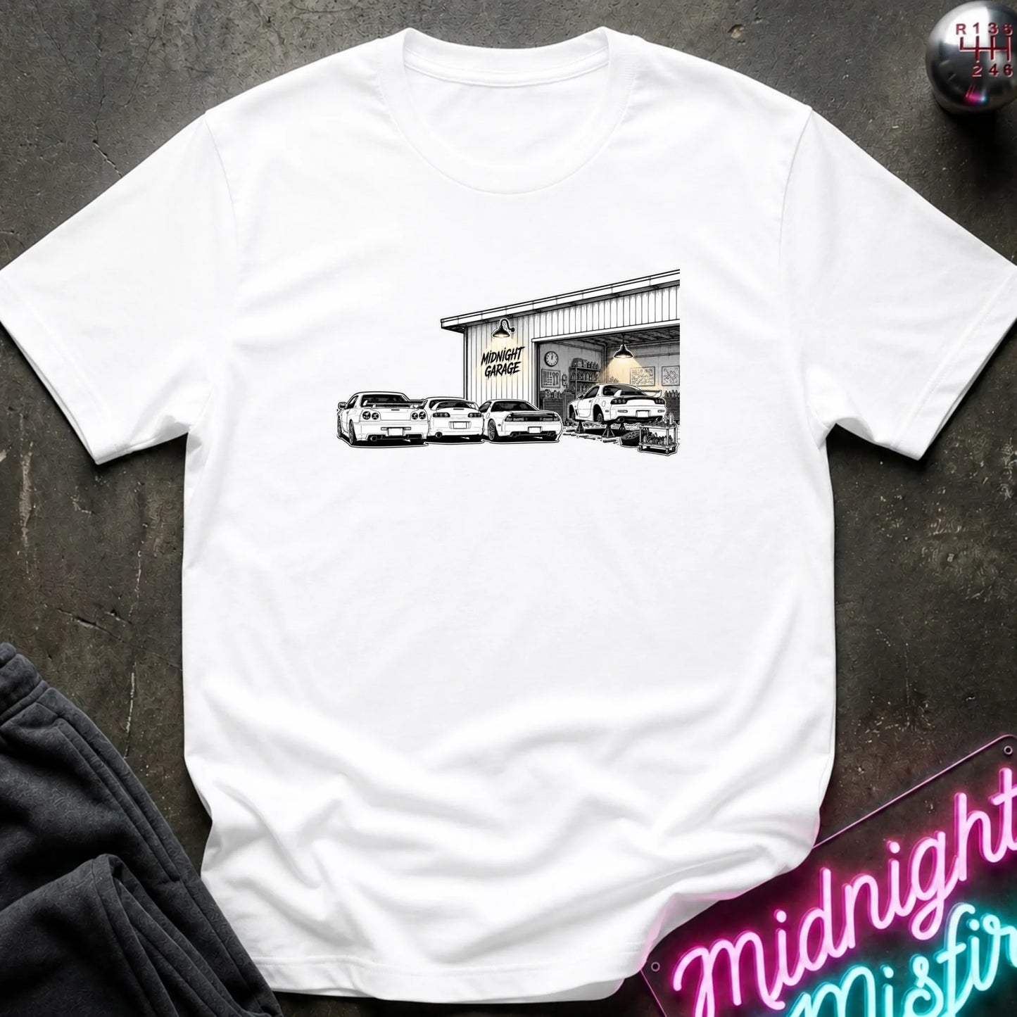 A white short-sleeve t-shirt laid flat on a dark textured surface, featuring a black and white graphic print of a garage with several cars in front. 
